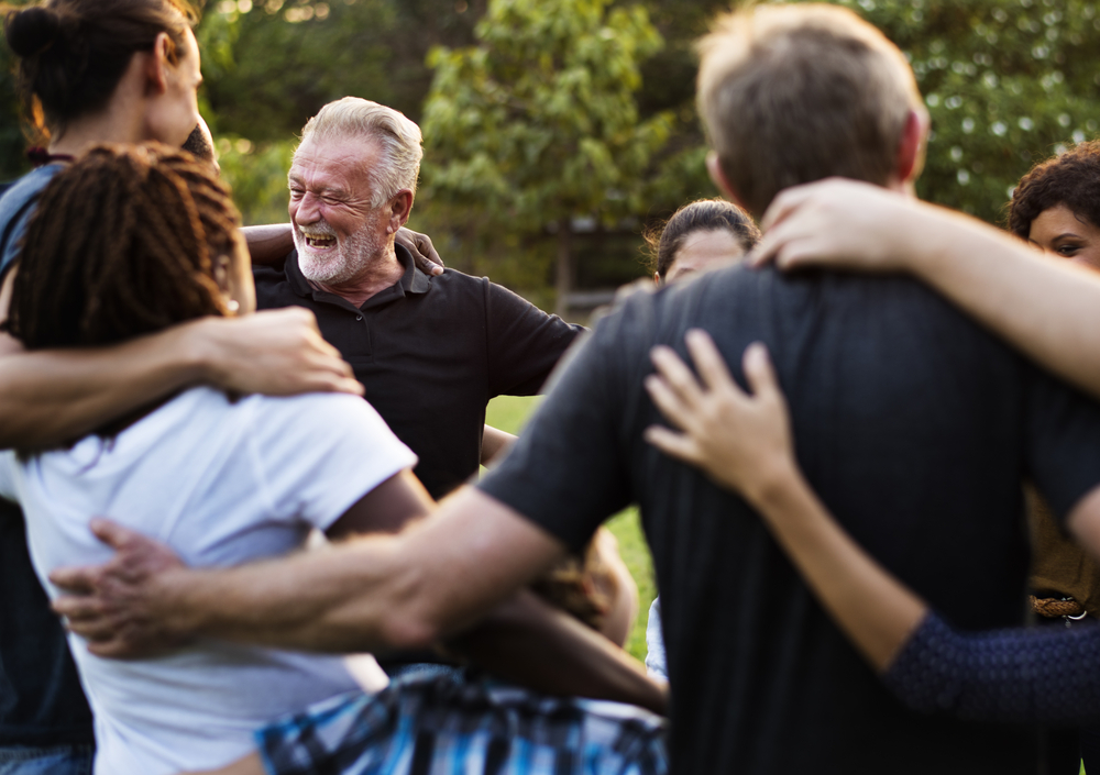 A diverse group of adults huddle together, embracing each other with arms around shoulders. An older man with a grey beard in the center is laughing heartily, conveying joy and connection. The image suggests strong community support and camaraderie.