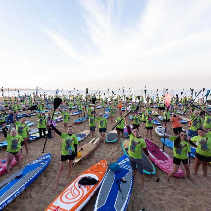 A large group of people, many in matching green shirts, gather on a sandy beach with their stand-up paddleboards. Many are smiling and holding their paddles triumphantly in the air, looking at the camera. The image conveys a sense of community and energetic participation in an outdoor event.