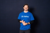 Rob Mills, an Australian singer and actor, smiles while wearing a blue Lifeline t-shirt over a black long-sleeved shirt. He has dark, wavy hair, a beard, and a watch on his left wrist. His hands are clasped together in front of him, and he stands against a dark blue background.