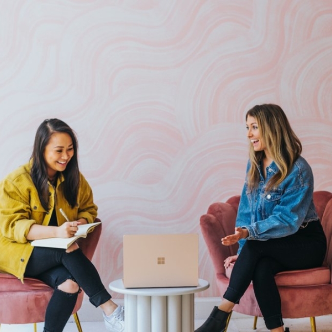 Two women sitting on chairs, smiling and talking while working together with a laptop on a small table between them