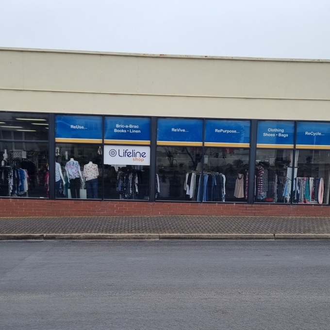 A Lifeline thrift shop with large storefront windows displaying clothing racks and mannequins. Blue banners stretch across the top of the windows, listing categories like "ReUse," "Bric-a-Brac Books Linen," "ReVive," "RePurpose," "Clothing Shoes Bags," and "ReCycle." The building has a light beige upper facade and a brick lower section. A black A-frame sign on the sidewalk reads "NOW OPEN." A red car is parked to the left, and a black SUV is visible in a parking area to the right, behind a blue barrier.