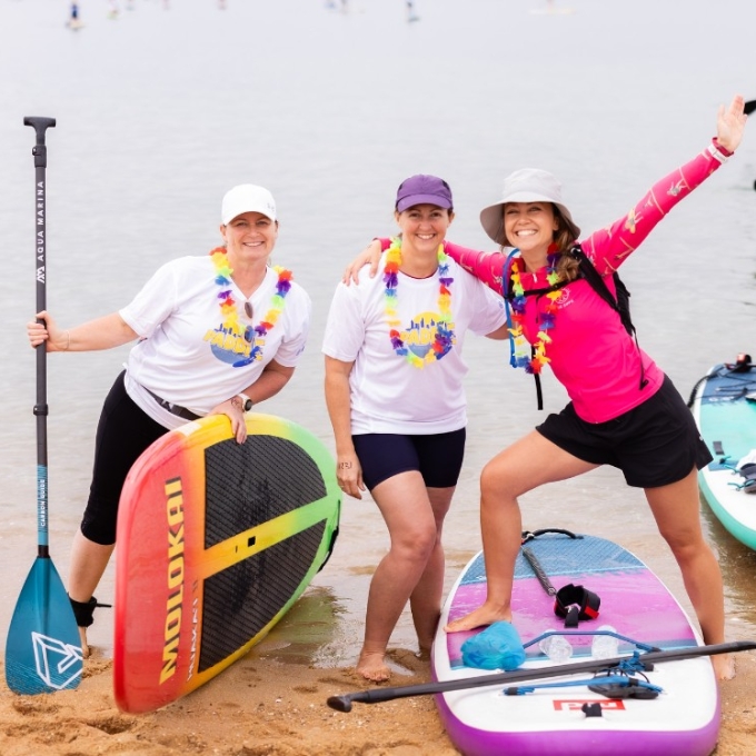 Three women in colourful leis smile on a beach next to their stand-up paddleboards. The woman on the left, in a white cap and T-shirt, holds a paddle. The middle woman, also in a white T-shirt and a purple cap, has her arm around the third woman. The third woman, in a pink long-sleeved top and a light-coloured hat, stands with one foot on a paddleboard and her arms raised in a cheerful pose. Other paddleboarders are visible in the water behind them.
