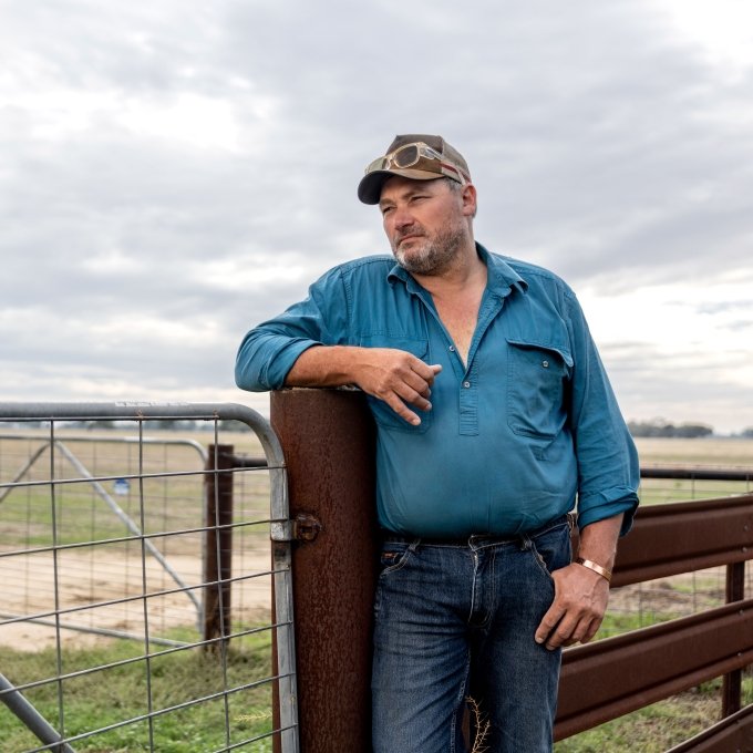 A middle aged Australian man in a rural setting, one arm learning on a gate, looking off into the distance.