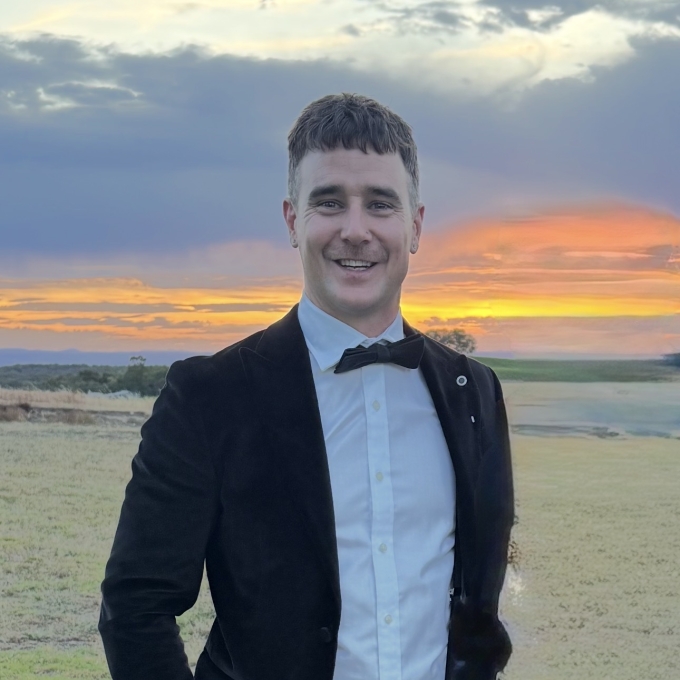 Mark smiling in a black suit and bow tie, standing outdoors at sunset.