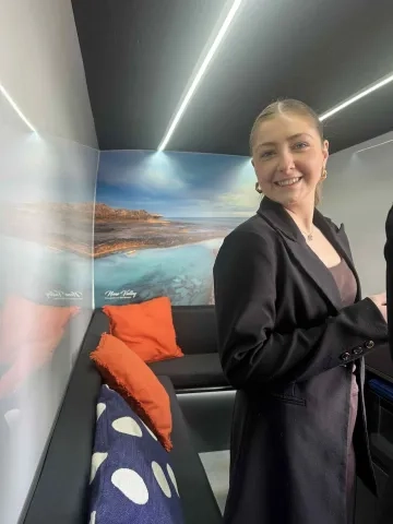 A smiling woman in a black blazer and brown top with gold hoop earrings and a silver necklace stands in a modern lounge area. Behind her, a panoramic mural depicts a serene coastal landscape with rocky shores meeting a clear turquoise sea under a bright sky. In the foreground, a black bench seat is adorned with two textured orange pillows and one blue pillow with white irregular spots. The ceiling features sleek white and dark gray panels with integrated strip lighting.