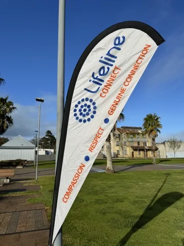 A white flag with "Lifeline," a circle of blue dots, "CONNECT," "GENUINE CONNECTION," "RESPECT," and "COMPASSION" in blue and orange text, angled against a clear blue sky. A grassy field, palm trees, and an old building are visible in the background.
