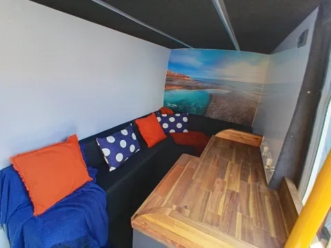 A compact seating area with a dark blue bench against a white wall. The bench is adorned with an orange throw blanket, two solid orange pillows, and two blue pillows with white polka dots. A colorful wall mural depicting a rocky seascape with turquoise water fills the far corner. Adjacent to the bench is a built-in wooden countertop with a butcher block pattern.